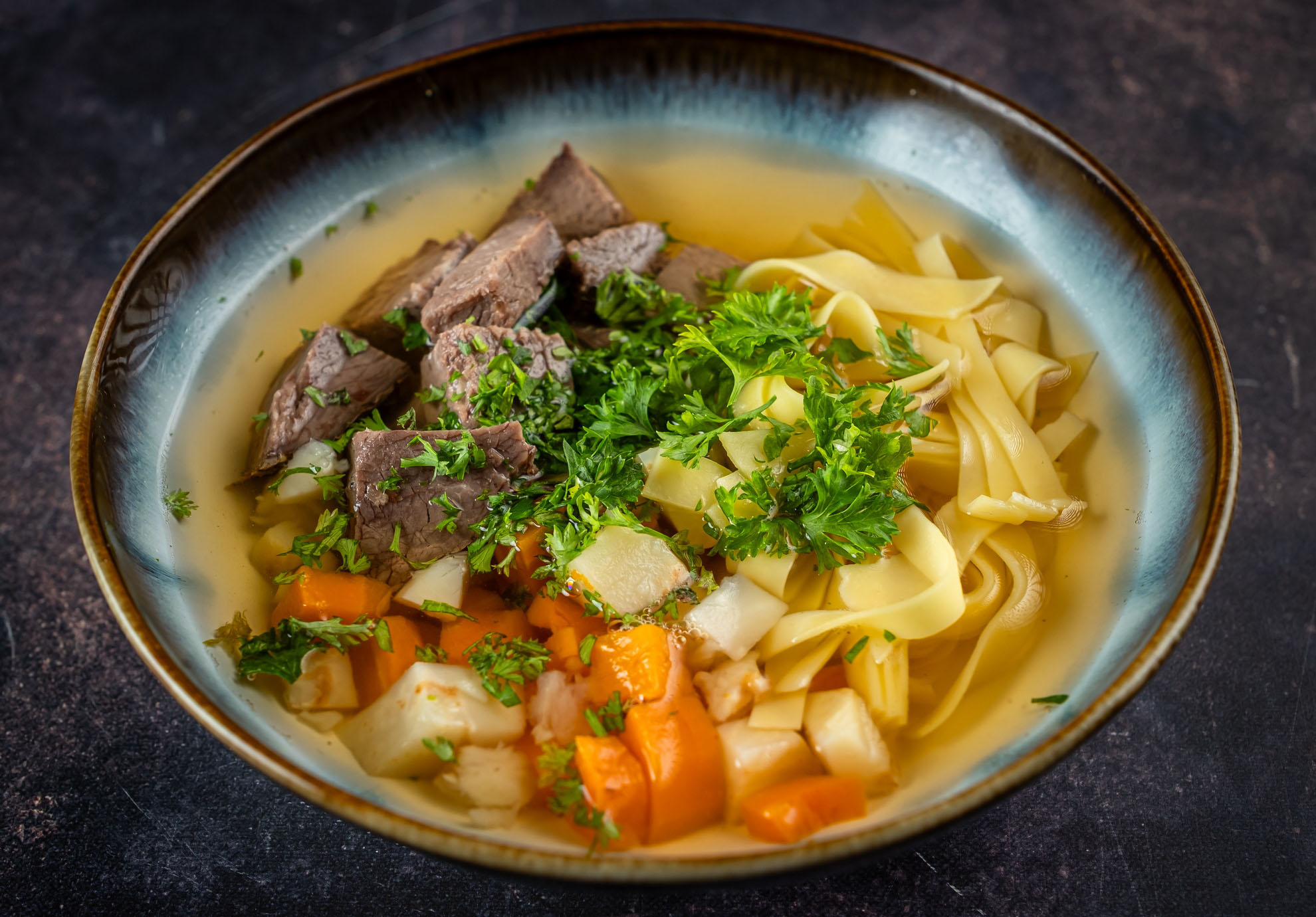 Eine Schüssel herzhafter Rindfleisch-Nudelsuppe mit zarten Rindfleischstücken, geschnittenen Karotten, gewürfelten Kartoffeln, Eiernudeln und frischer Petersilie, vor einem dunklen Hintergrund.